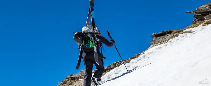 A skier with equipment on its back walks uphill on a snowy mountain.