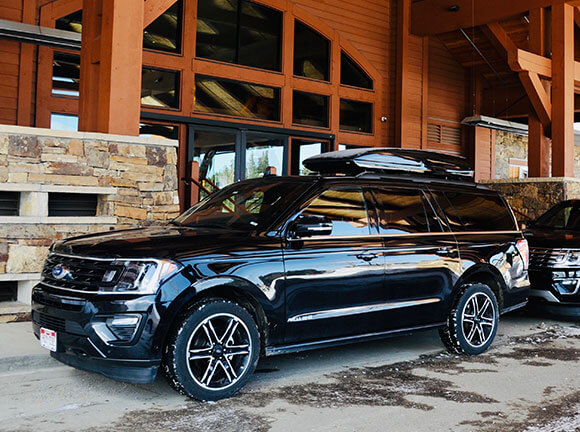 Two black SUVs and wooden house background.